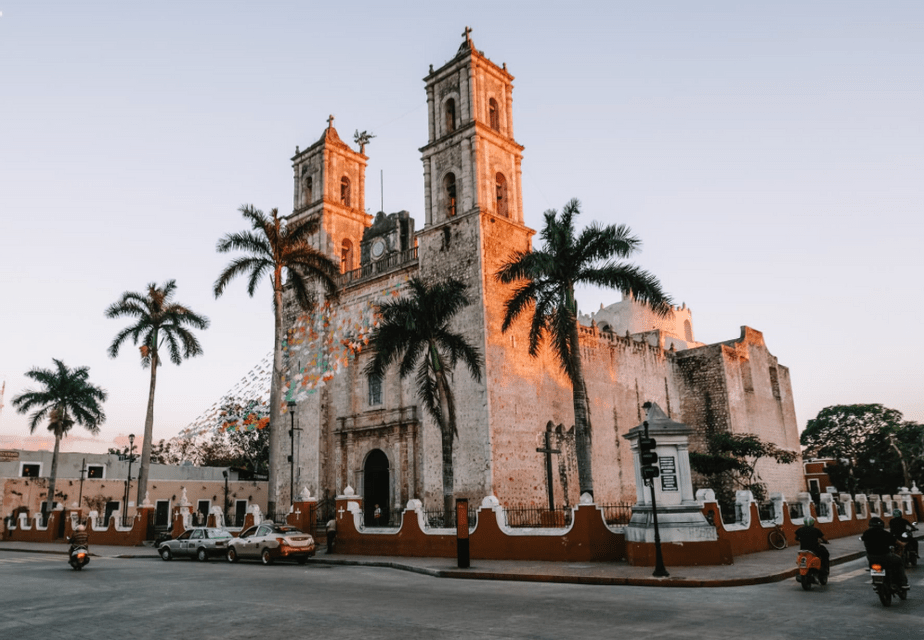 A large stone church with two bell towers glows in the sunset light on a street corner, surrounded by palm trees and passing traffic.