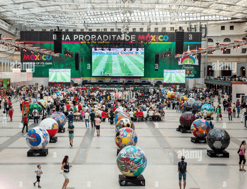 A large crowd gathers in an exhibition hall to watch a soccer match on big screens, with oversized, decorated soccer balls in the foreground.