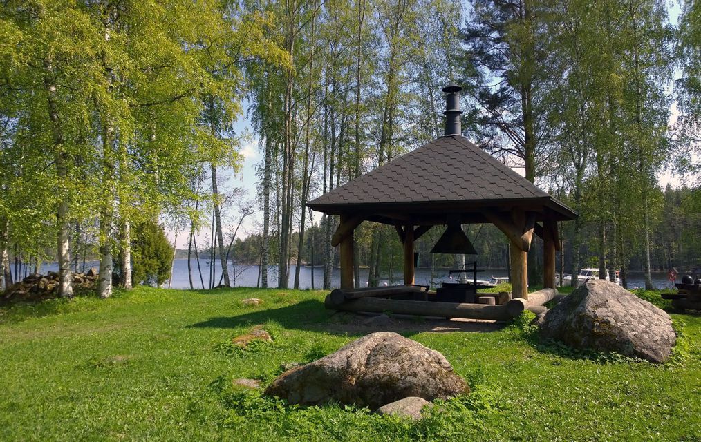 Ein hölzerner Grillpavillon mit Ziegeldach steht auf einer grünen Wiese neben einem von Birken umgebenen See.
