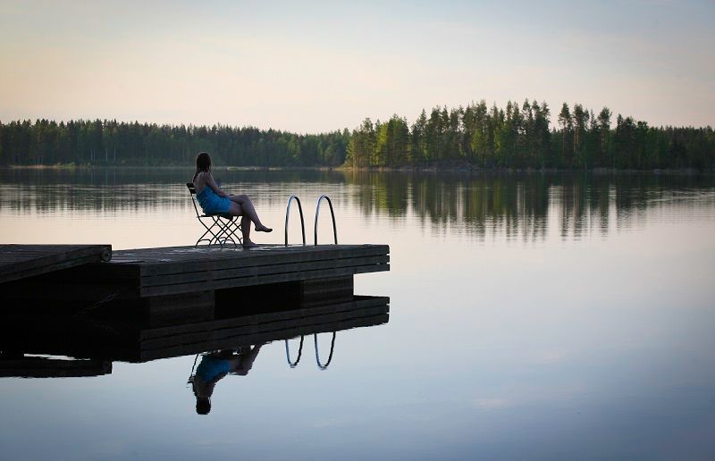 Eine Person sitzt auf einem Stuhl am Ende eines Holzstegs und blickt auf einen ruhigen See, der eine ferne, bewaldete Küstenlinie widerspiegelt.