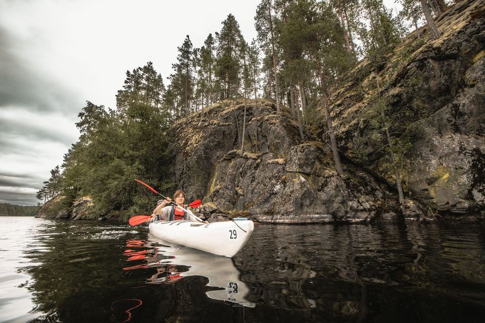 Eine Person paddelt in einem weißen Kajak auf dunklem Wasser neben einer felsigen, bewaldeten Küste unter einem bewölkten Himmel.