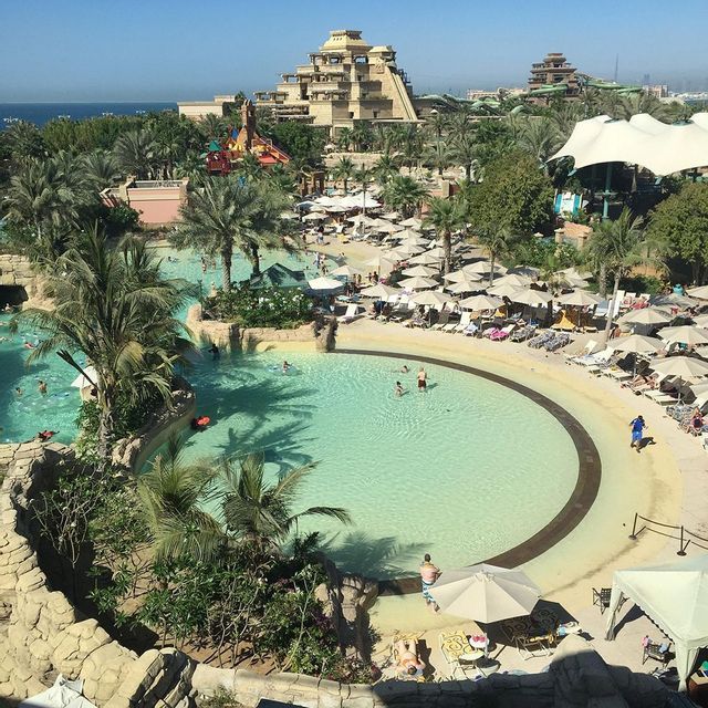 Ein weitläufiger Wasserpark, gefüllt mit Menschen, die in Pools schwimmen, umgeben von Palmen, Sonnenliegen und großen Resortgebäuden.
