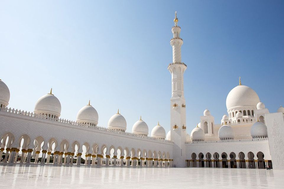 Eine große weiße Moschee mit mehreren Kuppeln, einem Minarett und einem Arkadenhof steht unter einem klaren blauen Himmel.