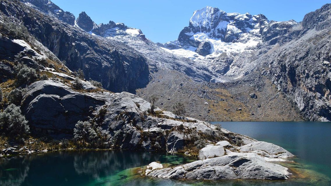 Un limpido lago alpino turchese con sponde rocciose giace ai piedi di imponenti montagne innevate sotto un cielo azzurro.