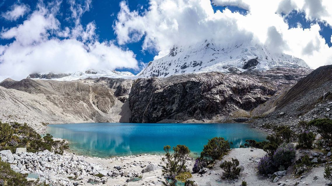 Un lago alpino turchese e vivace giace ai piedi di una catena montuosa rocciosa e innevata, sotto un cielo azzurro parzialmente nuvoloso.