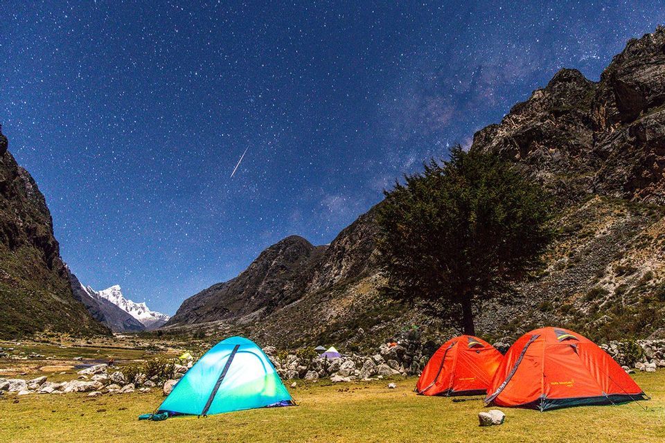 Tende da campeggio colorate sono montate in una valle di montagna erbosa sotto un cielo notturno sereno e stellato, con una stella cadente visibile.