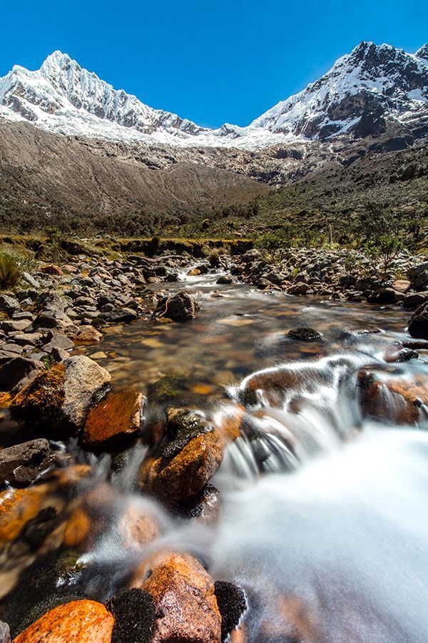 Un ruscello roccioso con acqua sfocata dal movimento scorre in una valle sotto montagne innevate, sotto un cielo azzurro.