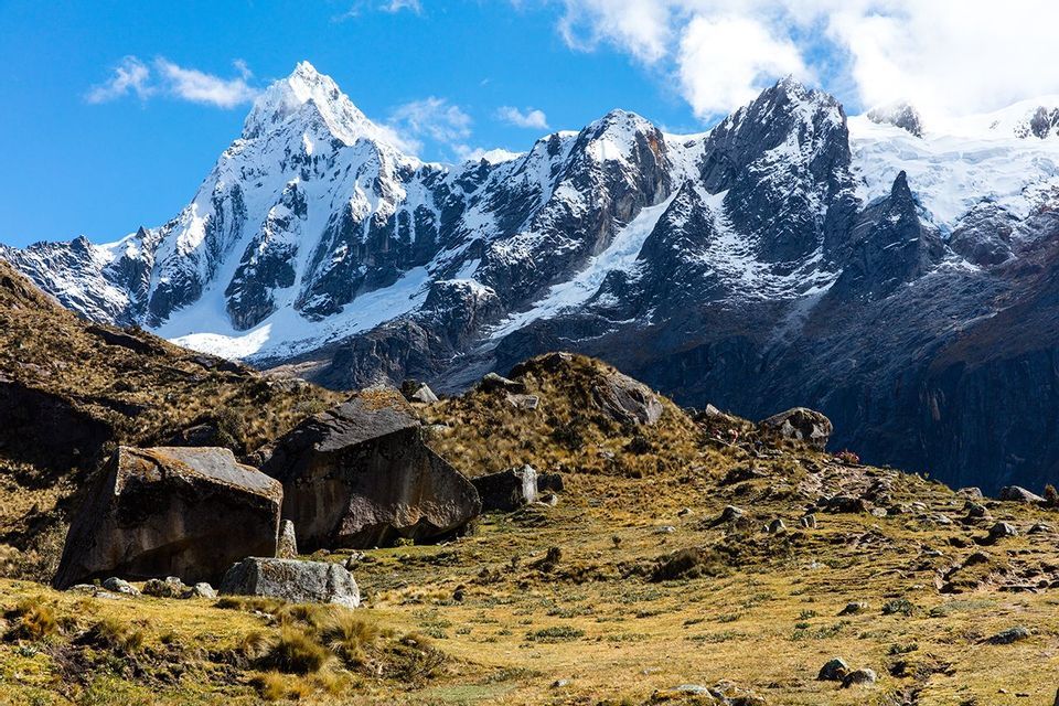 Imponenti montagne innevate dominano un paesaggio roccioso baciato dal sole, costellato di grandi massi e erba secca.