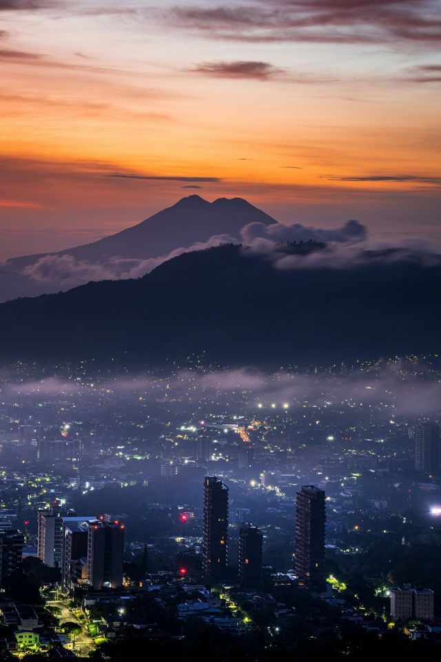 Una città con luci sfavillanti al tramonto, adagiata in una valle con montagne e un vulcano stagliati contro un cielo arancione.