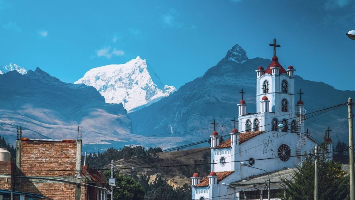 Una chiesa bianca dai tetti di tegole rosse si erge in una cittadina, con lo sfondo di un'imponente catena montuosa innevata sotto un cielo azzurro brillante.