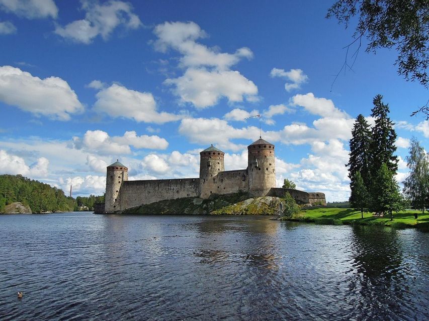 Eine mittelalterliche Steinburg mit mehreren Rundtürmen thront auf einer felsigen Küste neben einem großen Gewässer unter blauem Himmel mit flauschigen Wolken.