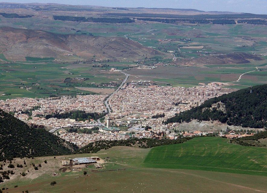 Una veduta aerea di una città tentacolare situata in una valle, circondata da dolci colline verdi e marroni e foreste.