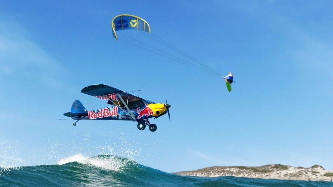 Un kitesurfer è in volo in un cielo azzurro e limpido, mentre un aereo a elica Red Bull vola basso sopra un'onda dell'oceano.