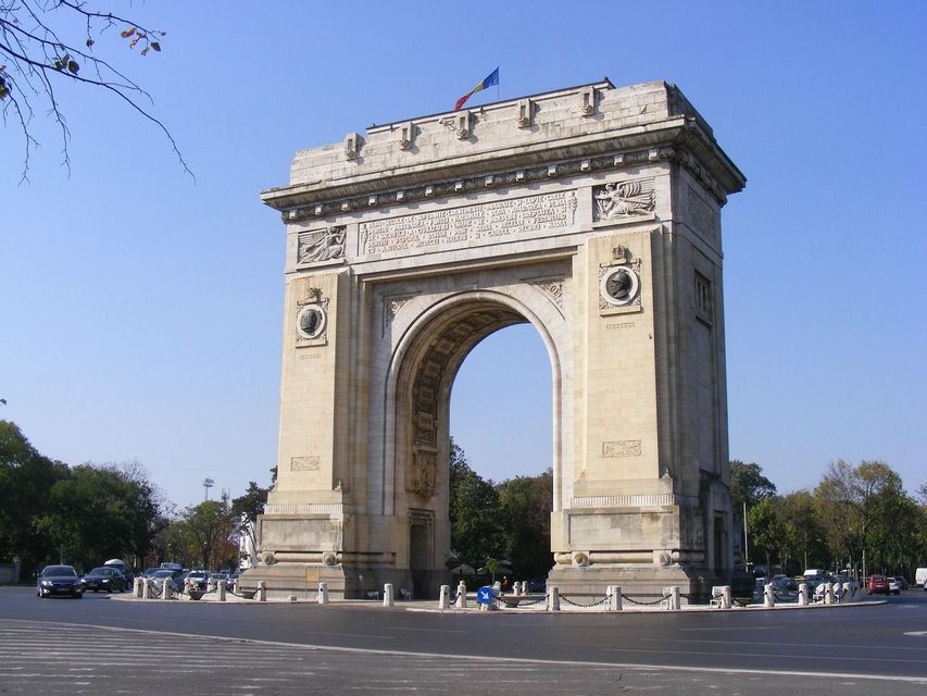 Un gran arco de triunfo de piedra se alza en el centro de una rotonda de tráfico con coches circulando a su alrededor bajo un cielo azul claro.