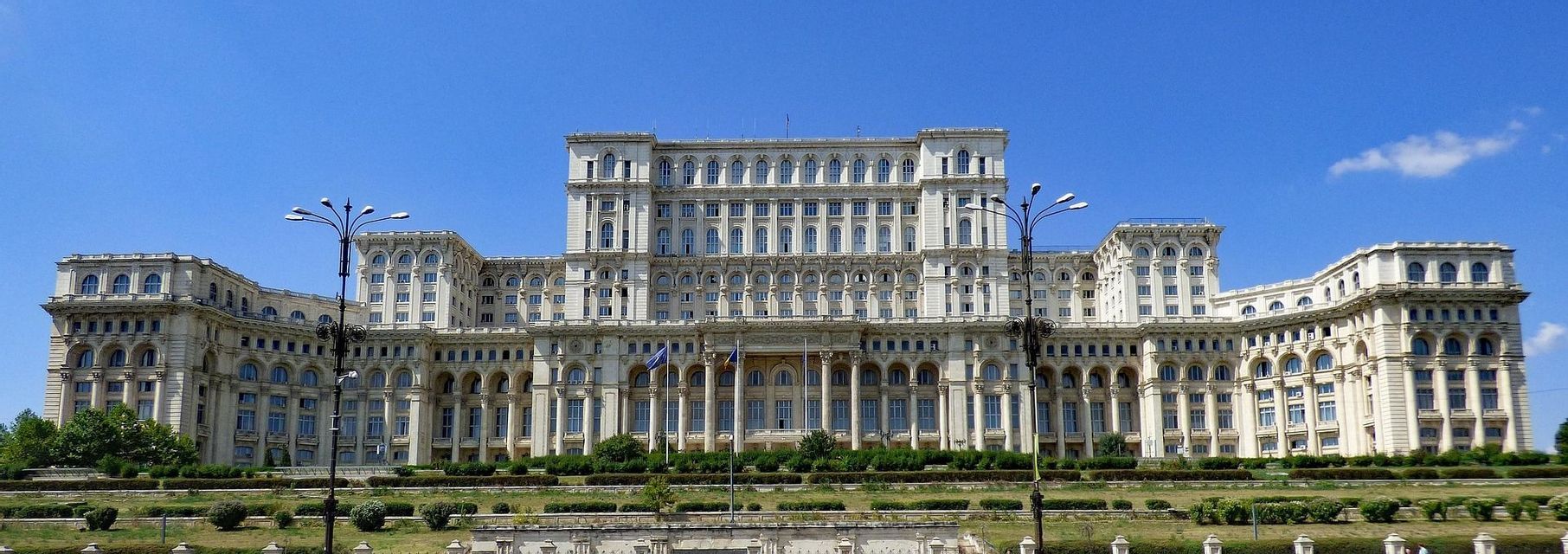 La fachada completa de un vasto y ornamentado edificio blanco con muchas ventanas se asienta detrás de un césped verde bajo un cielo azul claro.