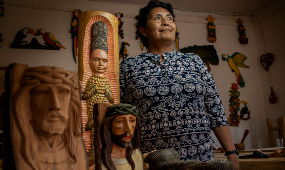 Un'artigiana nel suo laboratorio tra sculture in legno intagliate, come figure religiose e uccelli colorati.