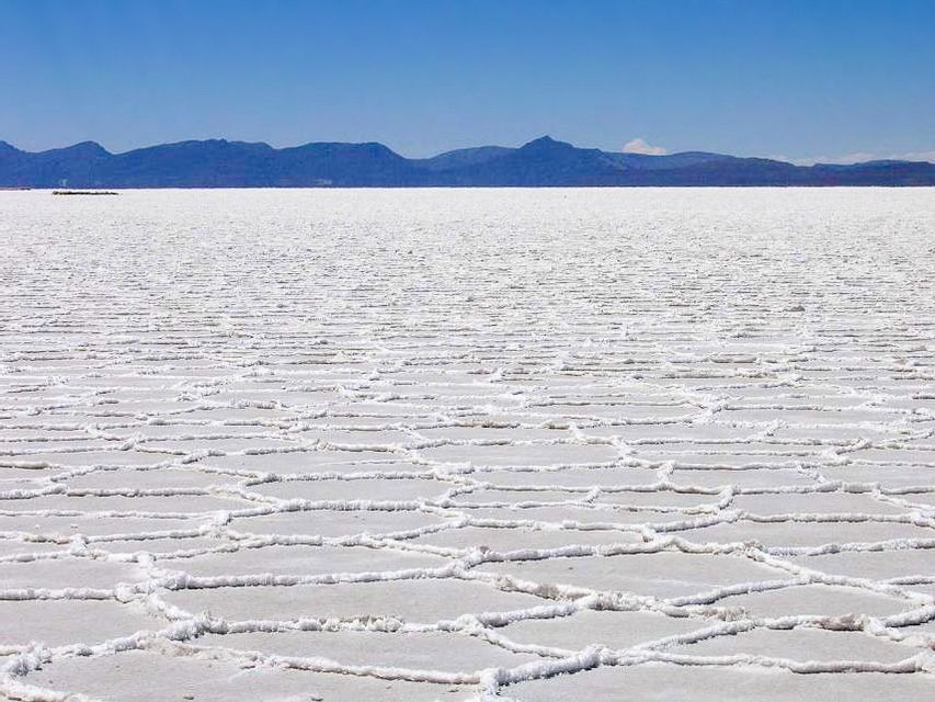 Una vasta distesa salina con naturali motivi esagonali che si estende verso montagne lontane sotto un cielo azzurro e limpido.