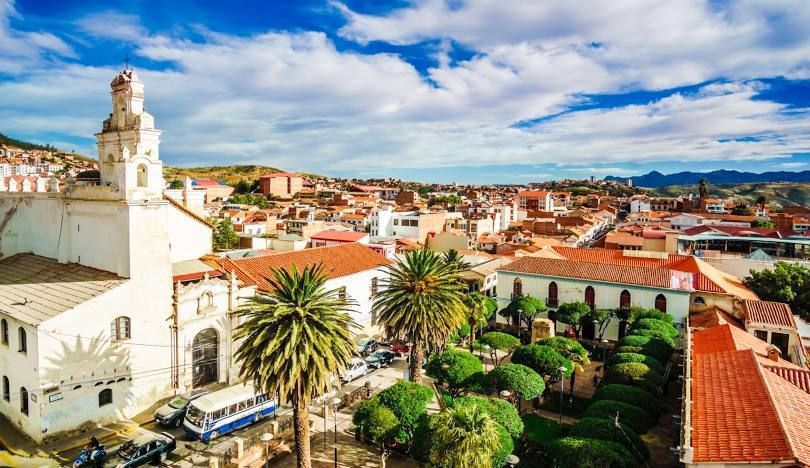 Veduta dall'alto di una città con una chiesa bianca, una piazza centrale con palme e tetti rossi sotto un cielo parzialmente nuvoloso.