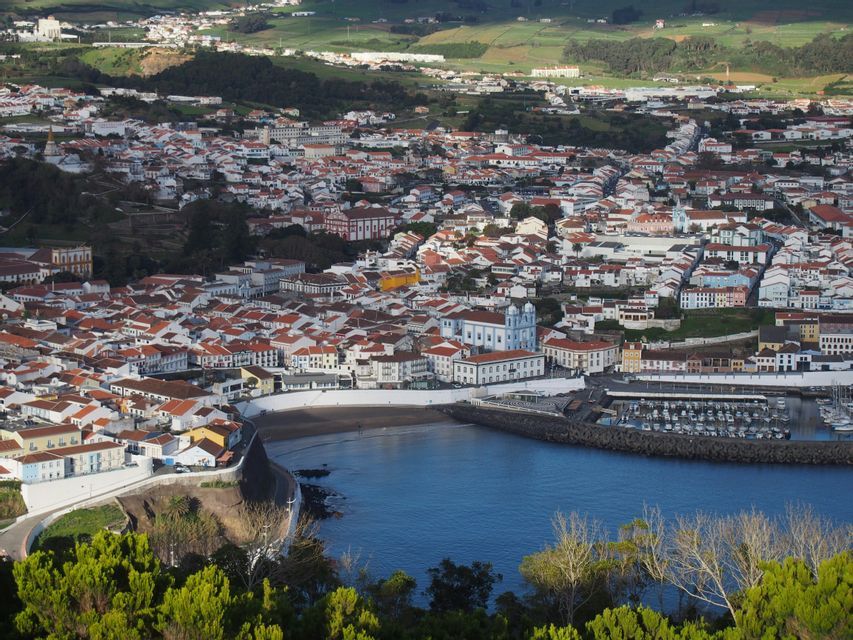 Una vista dall'alto di una cittadina costiera con edifici bianchi e tetti in terracotta che circondano una baia blu con un porto turistico.
