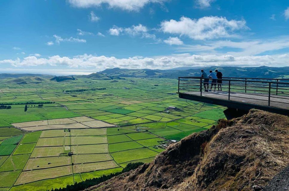 Un viaggio di gruppo WeRoad di tre persone su un punto panoramico con vista su un vasto paesaggio di campi verdi a mosaico sotto un cielo blu e nuvoloso.