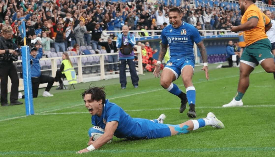 Un giocatore di rugby in divisa blu esulta dopo aver segnato una meta, con i compagni di squadra e la folla che lo acclama sullo sfondo dello stadio.
