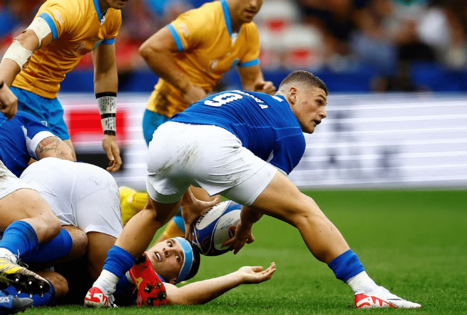 Un giocatore di rugby in maglia blu con il numero 9 passa la palla sopra un giocatore placcato sull'erba durante una partita.