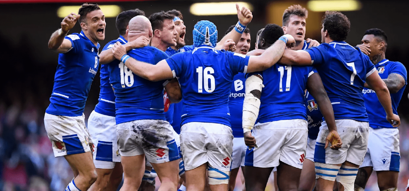 Un gruppo di giocatori di rugby con maglie blu e pantaloncini bianchi infangati festeggia in un abbraccio stretto sul campo, urlando e stringendosi.
