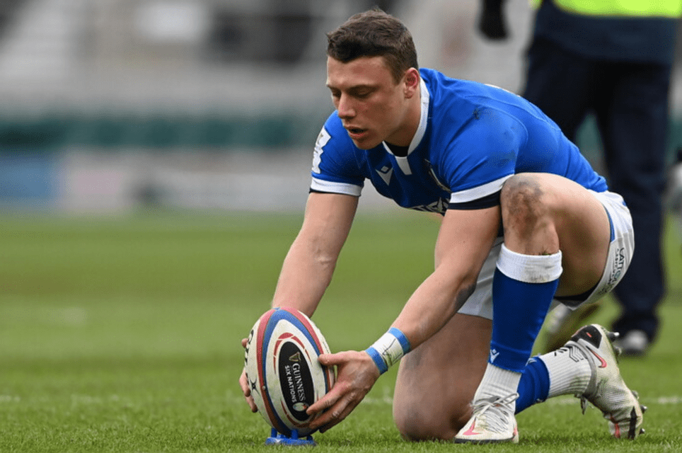 Un giocatore di rugby con una maglia blu si inginocchia su un campo erboso, posizionando una palla da rugby su un tee blu.