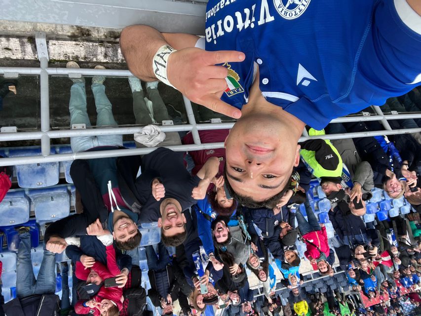 Un gruppo WeRoad in viaggio sorride per un selfie scattato da un uomo con una maglia blu sui sedili dello stadio.