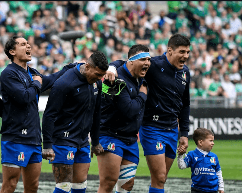 Quattro giocatori della nazionale italiana di rugby cantano sul campo, uno tiene per mano un bambino con la stessa maglia.