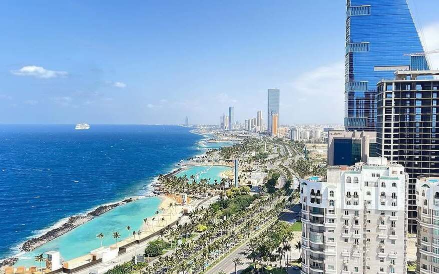 Una vista aerea di una moderna città costiera con grattacieli, spiagge orlate di palme e lagune turchesi accanto al mare blu profondo.