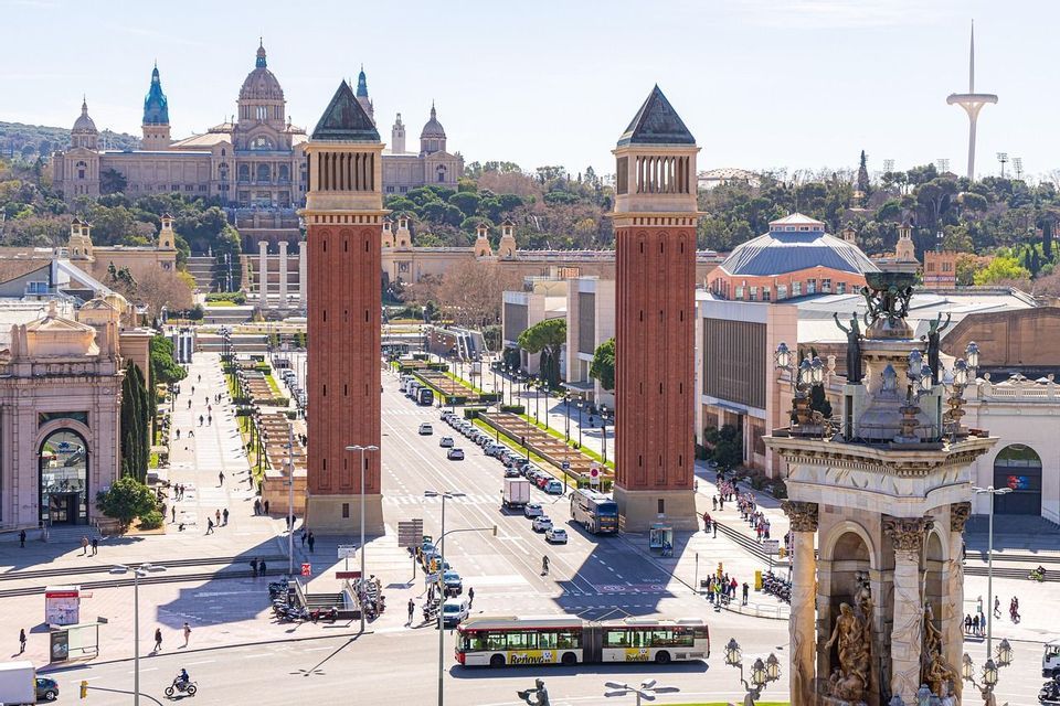 A sunny cityscape view of a wide avenue flanked by two large brick towers leading towards a palace on a hill.
