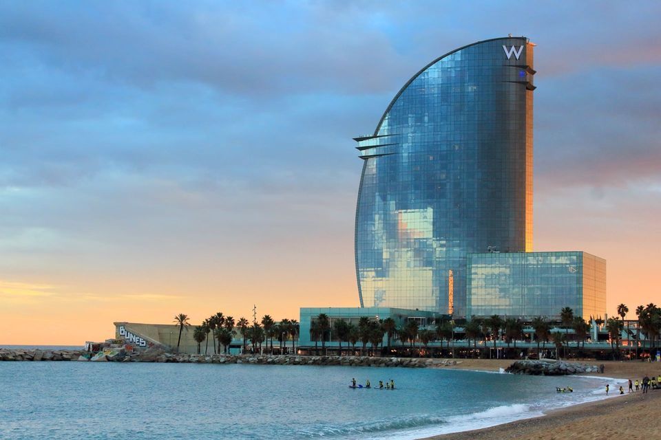 A modern sail-shaped glass building towers over a sandy beach at sunset, with palm trees and people on paddleboards in the water.