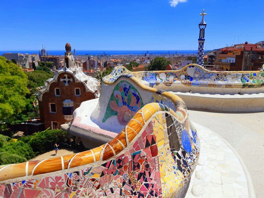 A colorful, curving mosaic bench on a terrace overlooking a cityscape and the sea under a clear blue sky.