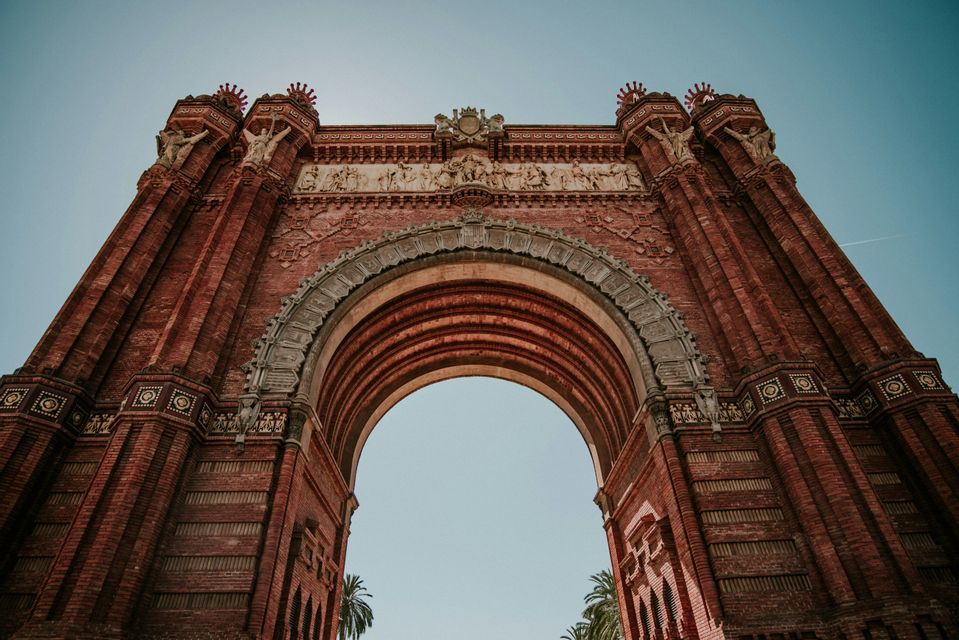 Una ripresa dal basso di un elaborato arco trionfale in mattoni rossi con sculture intricate, sullo sfondo di un cielo azzurro limpido.