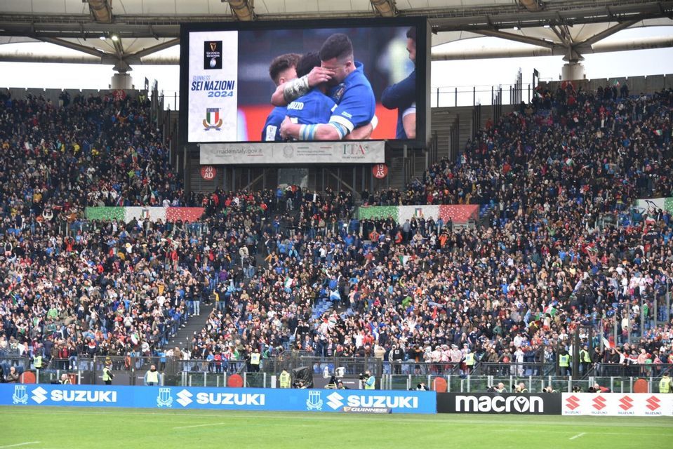 Una grande folla in uno stadio osserva un momento emozionante tra due giocatori di rugby italiani su un maxischermo durante il Sei Nazioni 2024.