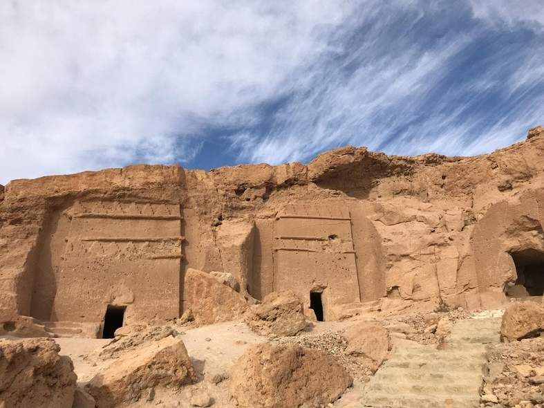 Antiche tombe con facciate rettangolari scavate in una scogliera rocciosa di arenaria sotto un cielo blu con nuvole sottili.