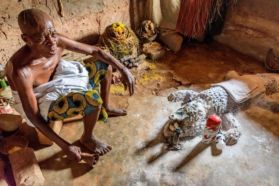 Un uomo anziano siede a torso nudo su uno sgabello basso all'interno di una capanna di fango, con un feticcio voodoo coperto di polvere sul pavimento accanto a lui.