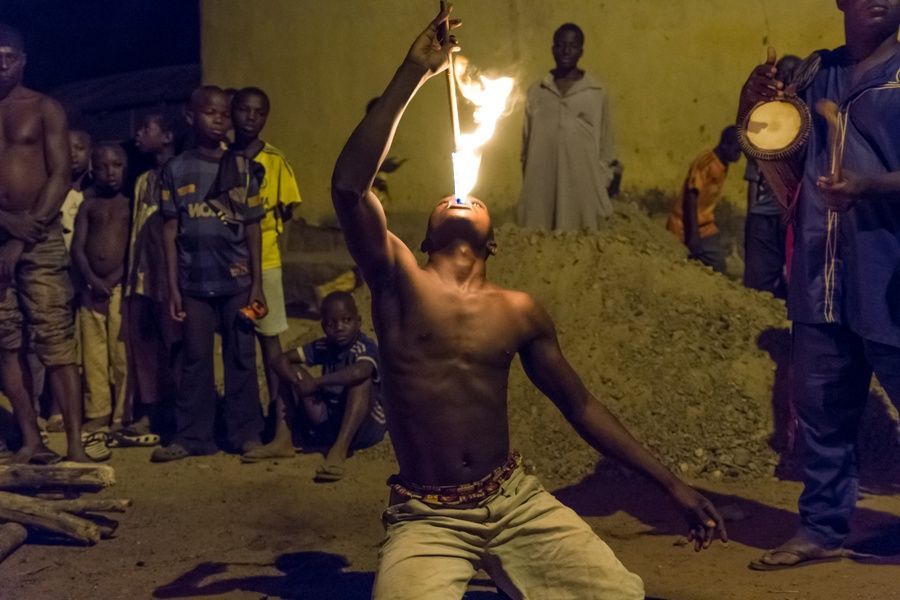 Un uomo a torso nudo si inginocchia a terra di notte, sputando fuoco da un bastone mentre una folla di persone guarda.