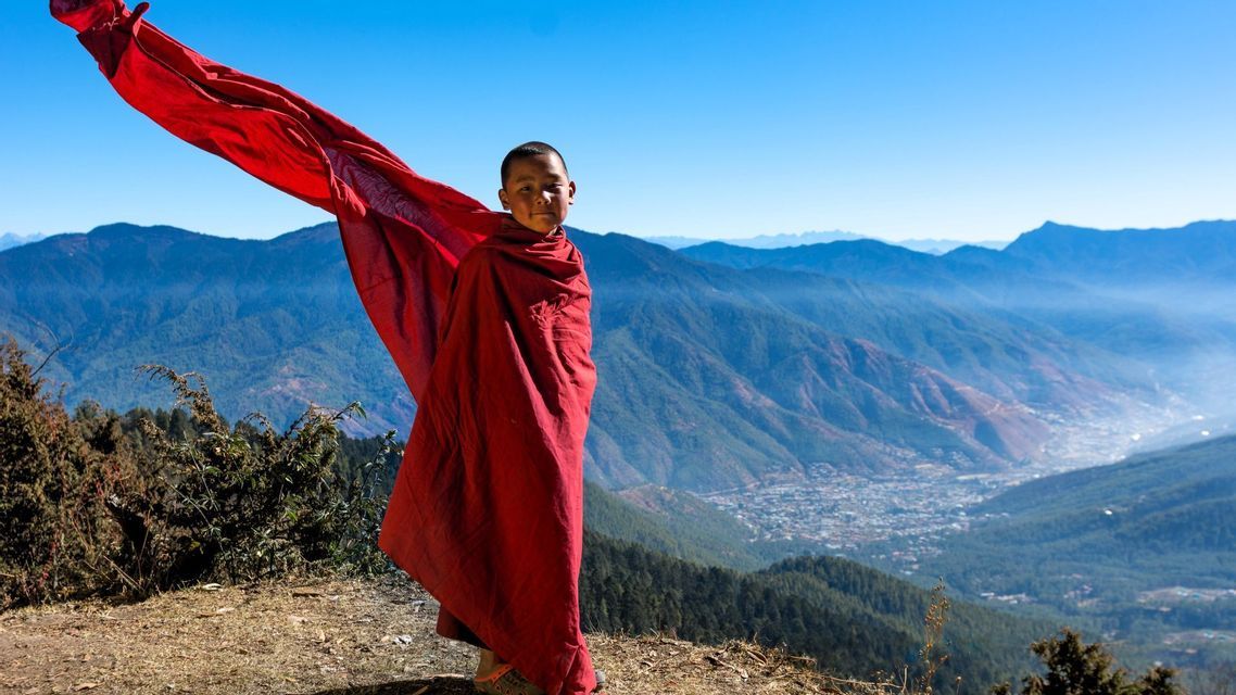 Ein junger buddhistischer Mönch steht auf einem Bergvorsprung, sein rotes Gewand flattert im Wind vor einer Kulisse aus Bergen und einem Tal.