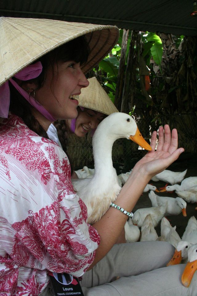 Una donna sorridente, durante un viaggio di gruppo WeRoad, con un cappello conico, nutre un'anatra bianca dalla sua mano, circondata da uno stormo.