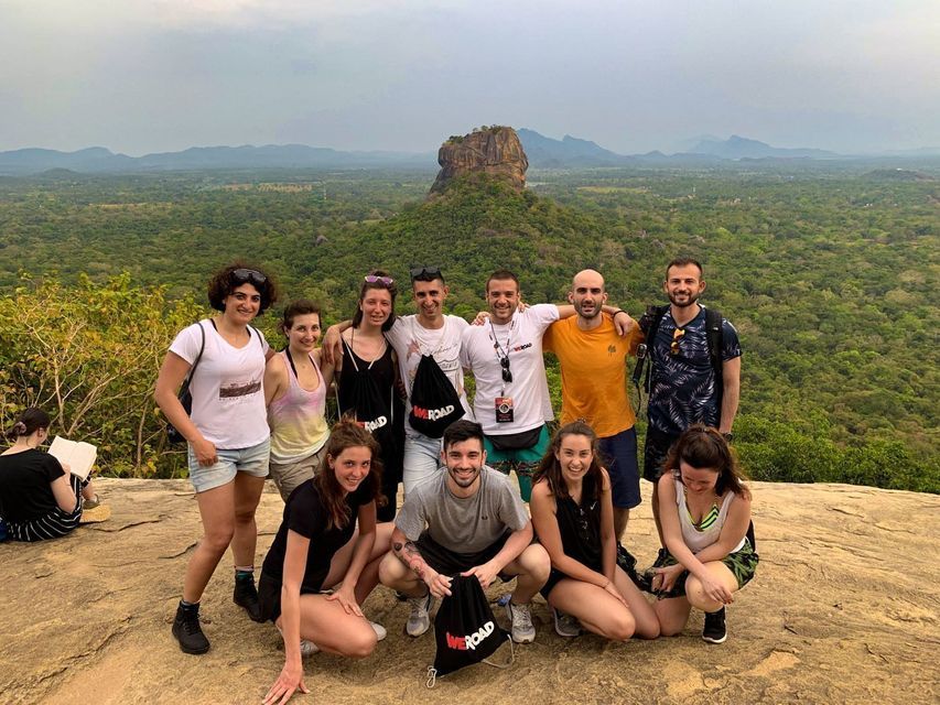 Un gruppo WeRoad in posa per una foto da un punto panoramico roccioso che si affaccia su una valle lussureggiante e una fortezza di roccia in lontananza.