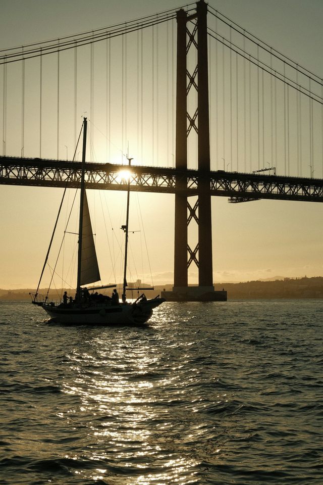 Un viaggio di gruppo WeRoad su una barca a vela stagliata contro il tramonto mentre naviga sotto un grande ponte sospeso.