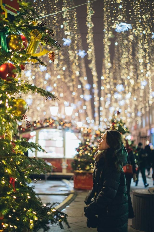 Una donna con un piumino nero guarda un albero di Natale decorato su una strada illuminata da luci festive di notte.