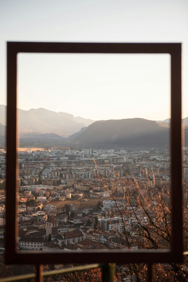 Un paesaggio urbano in una valle di montagna, visto attraverso una grande cornice rettangolare in primo piano, illuminato da calda luce solare.