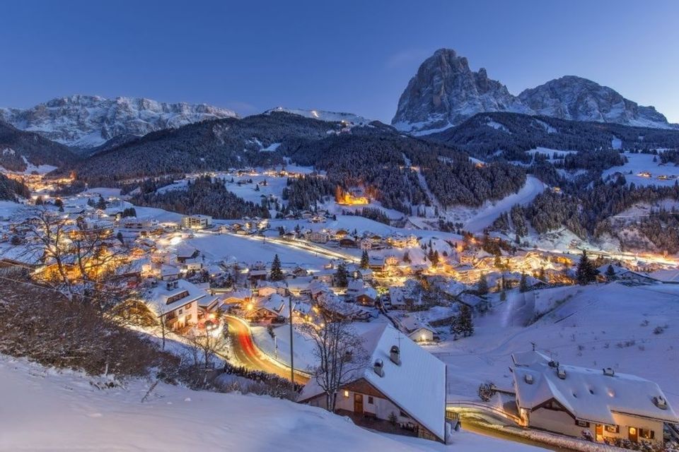 Un villaggio di montagna coperto di neve si illumina di luci calde al tramonto, con grandi cime rocciose sullo sfondo.