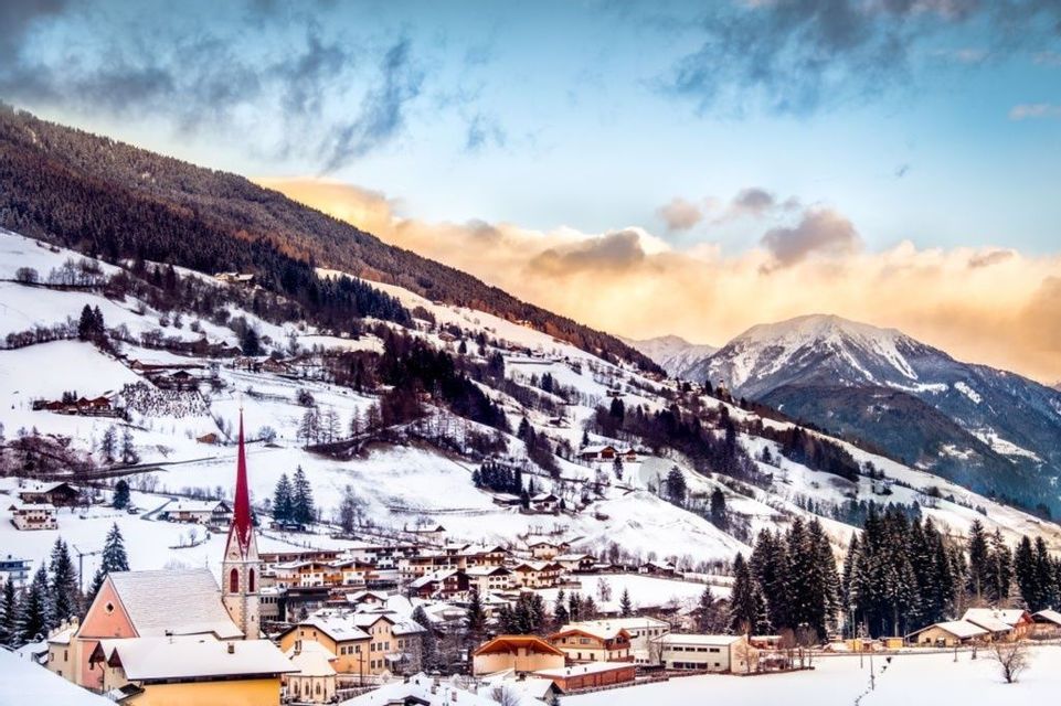 Un villaggio innevato con una chiesa dalla guglia rossa si adagia ai piedi di una catena montuosa boscosa, sotto un cielo nuvoloso al tramonto.