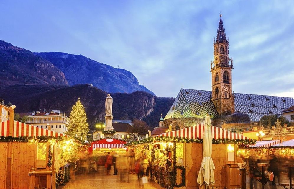 Un mercatino di Natale splendidamente illuminato in una piazza cittadina al tramonto, con una cattedrale storica e montagne sullo sfondo.