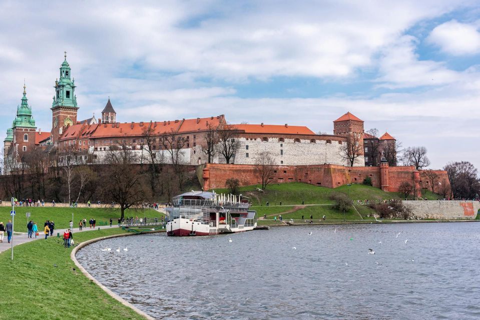 Un grande castello storico si erge su una verde collina che domina un ampio fiume, con una barca ormeggiata e persone che passeggiano lungo la riva.