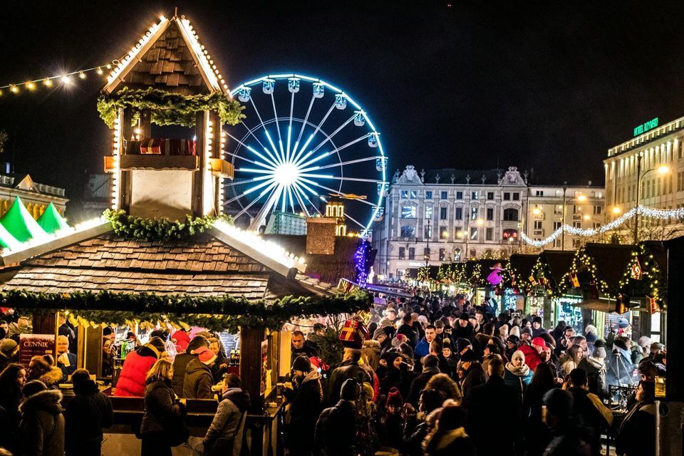 Un affollato mercatino di Natale notturno, con persone radunate alle bancarelle di legno e una grande ruota panoramica illuminata sullo sfondo.